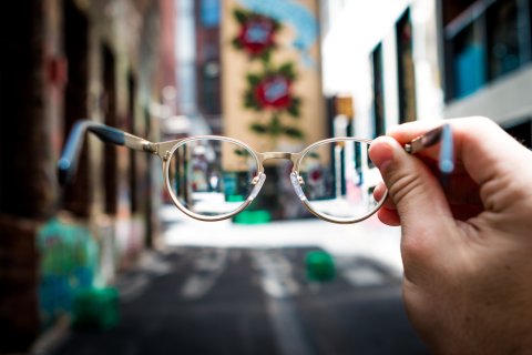 Blurred photo taken on a city street, in the foreground a pair of glasses, held by one hand