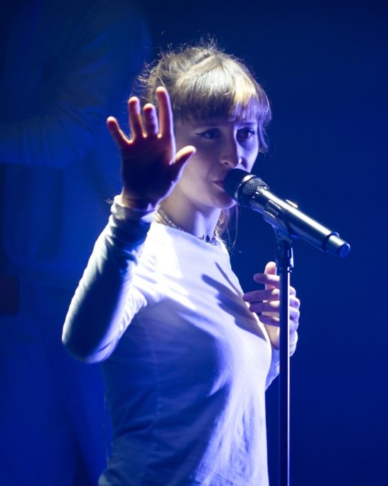 Chanteuse sur scène, main levée devant un microphone, éclairée en bleu.